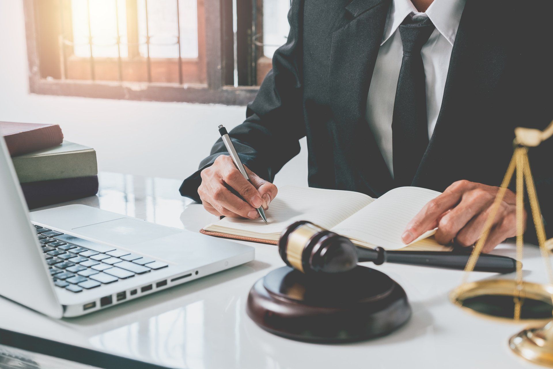 Lawyer writing at a desk with a gavel, laptop, and scales of justice