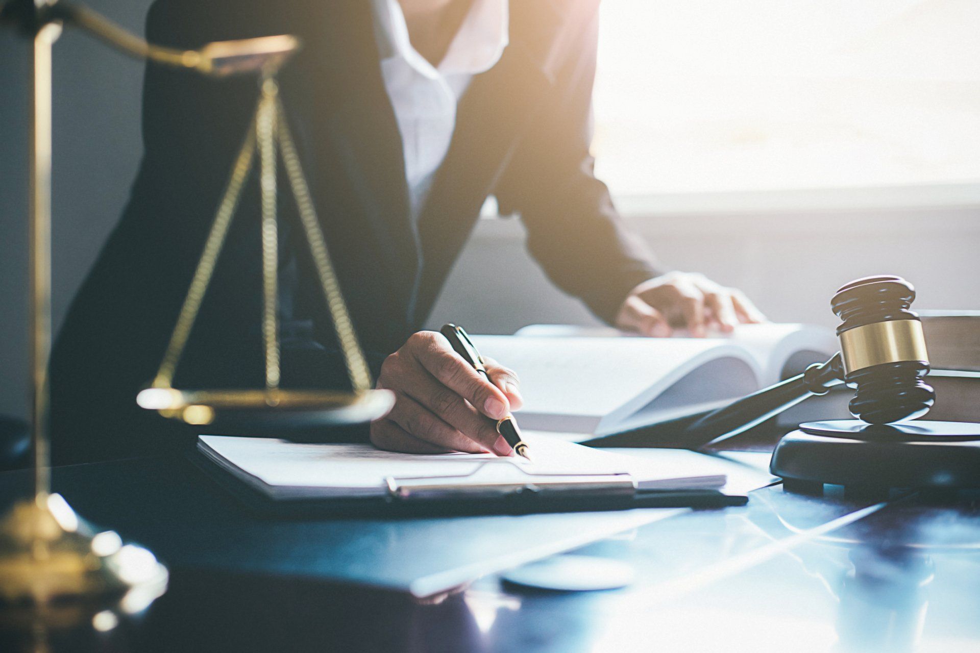 Lawyer signing documents at a desk with scales of justice and a gavel in a sunlit office