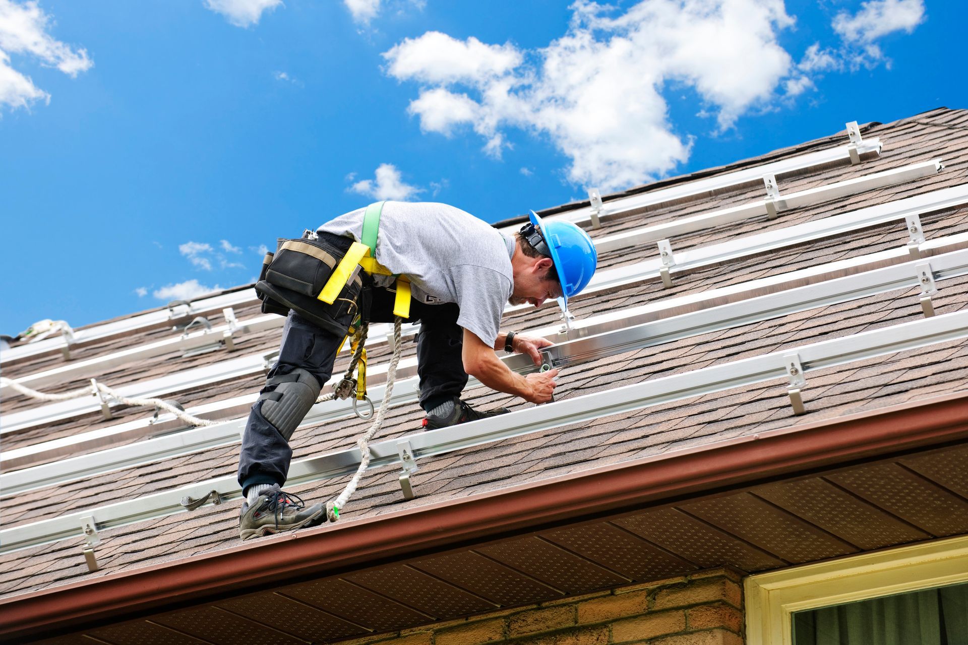 Commercial roof repair contractor securing metal brackets on shingle roof under blue sky.