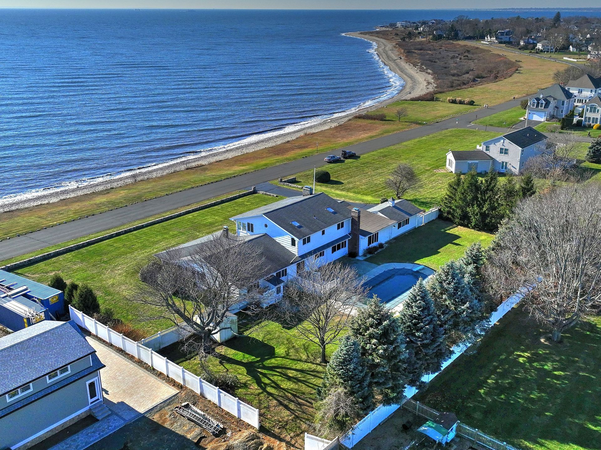 Aerial view of a coastal house with a pool, next to a beach and road, surrounded by lawn and trees.