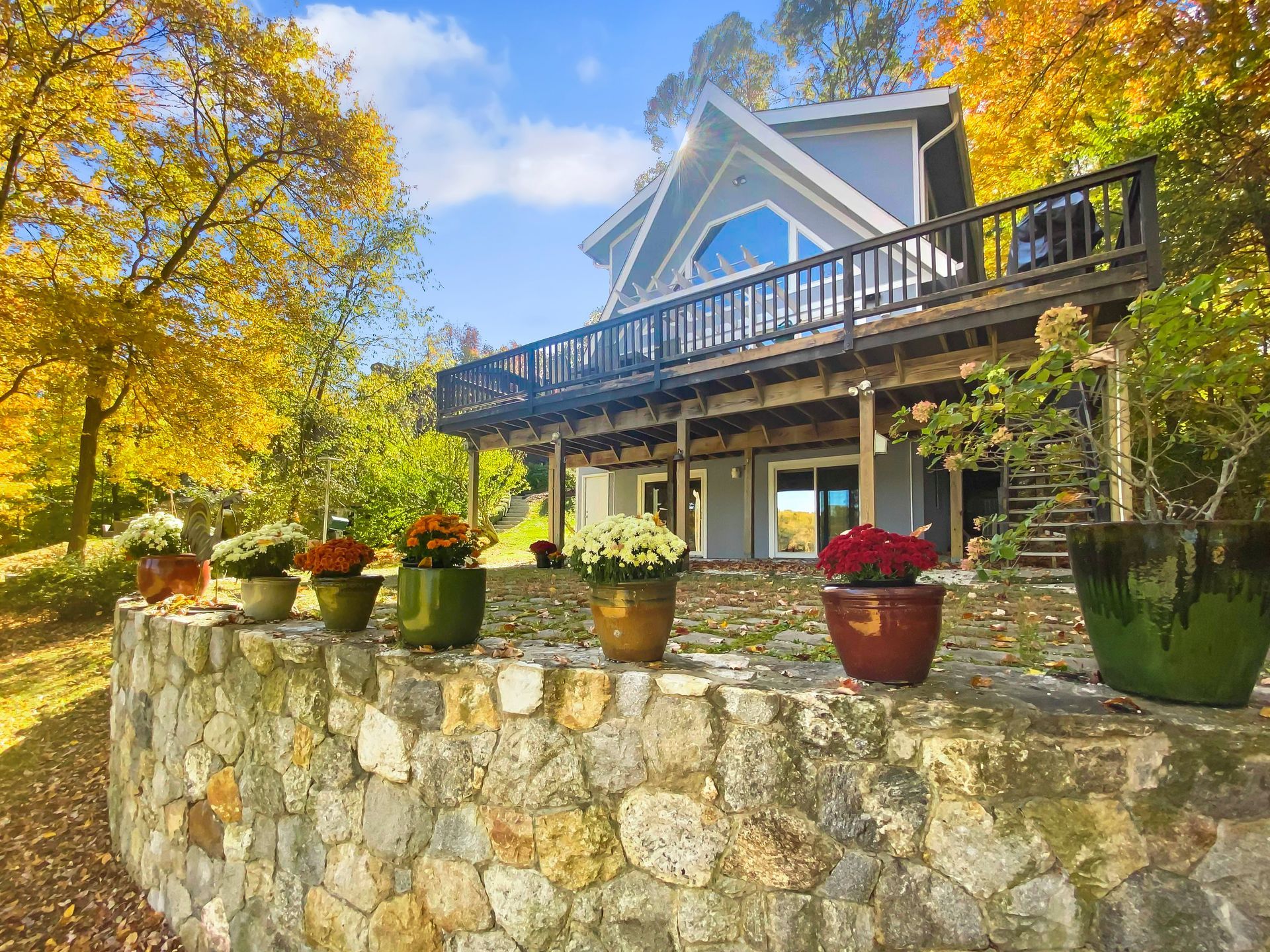 A two-story blue house with a wooden deck stands behind a stone retaining wall topped with potted flowers in autumn.