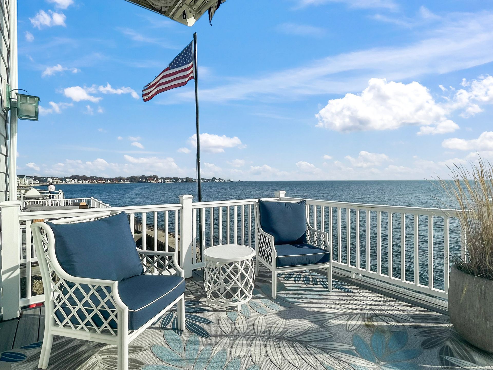 Two white lattice-style chairs with navy cushions on a deck overlooking a calm bay with an American flag under a blue sky.