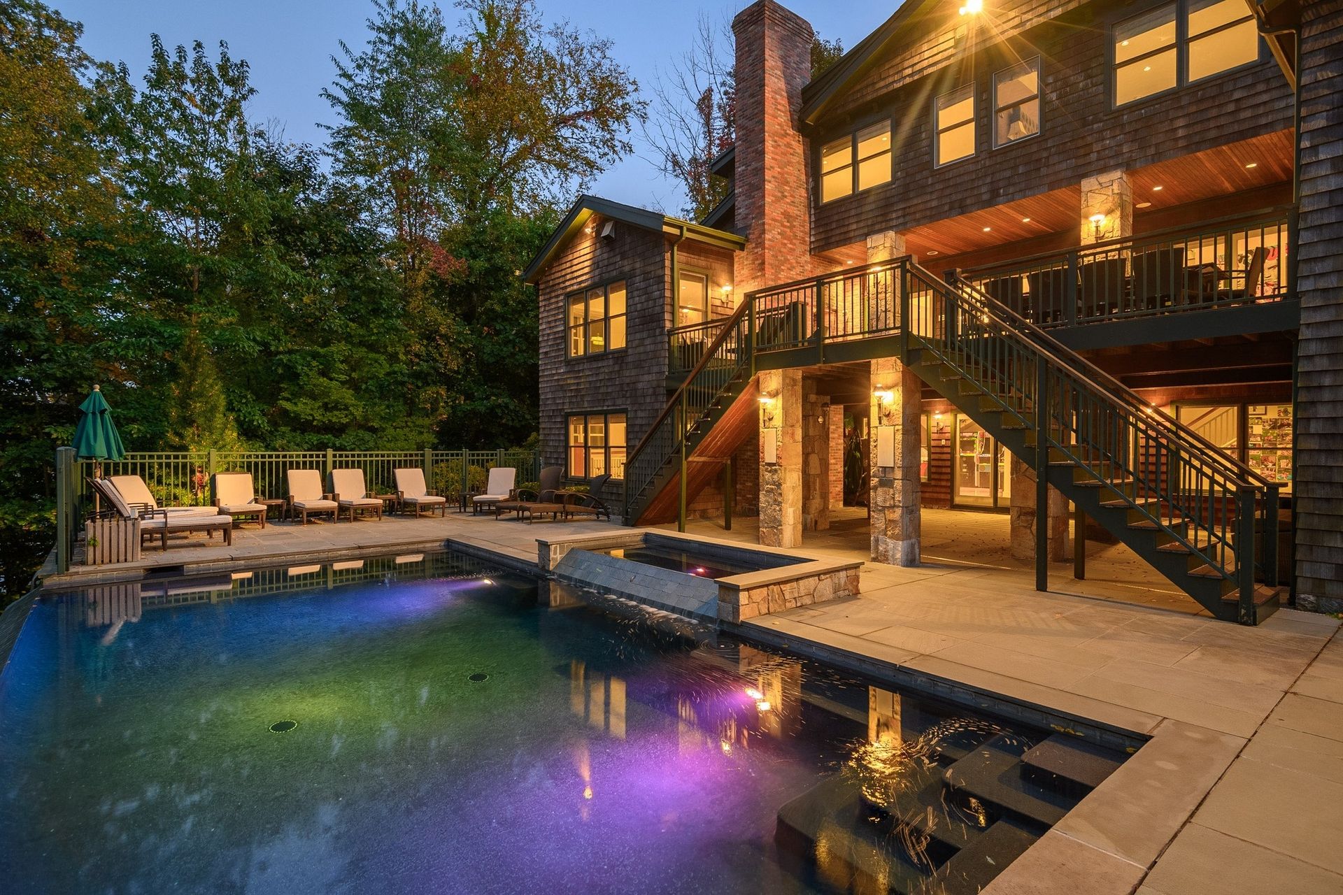 A multi-story stone home with an illuminated pool, patio chairs, and a grand staircase at dusk.