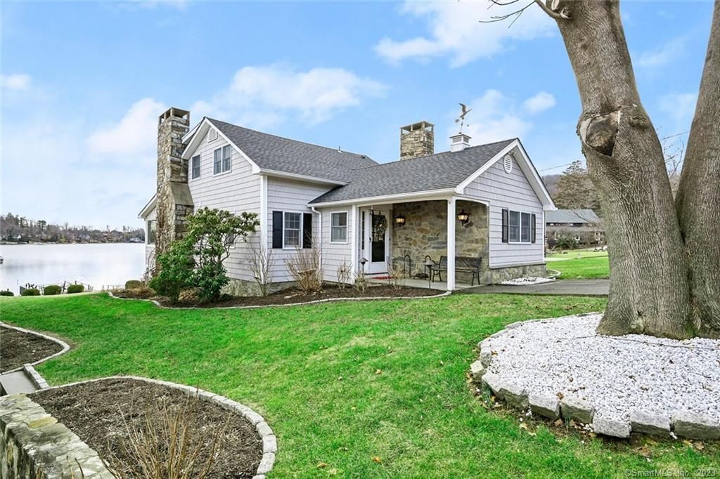 A white house with stone chimneys and siding sits on a green lawn by a lake, framed by a large tree in the foreground.