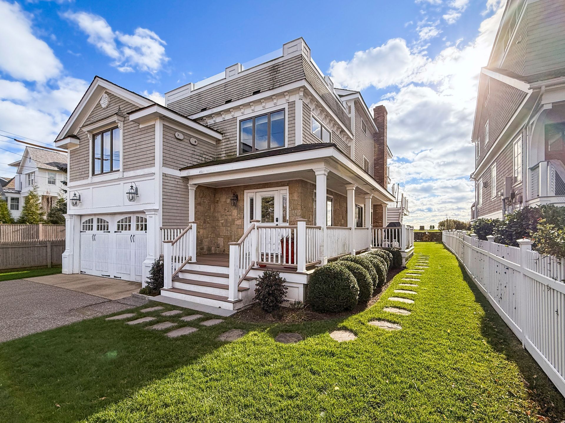 A two-story shingled house with a white wraparound porch, a front-facing garage, and a lush green lawn.