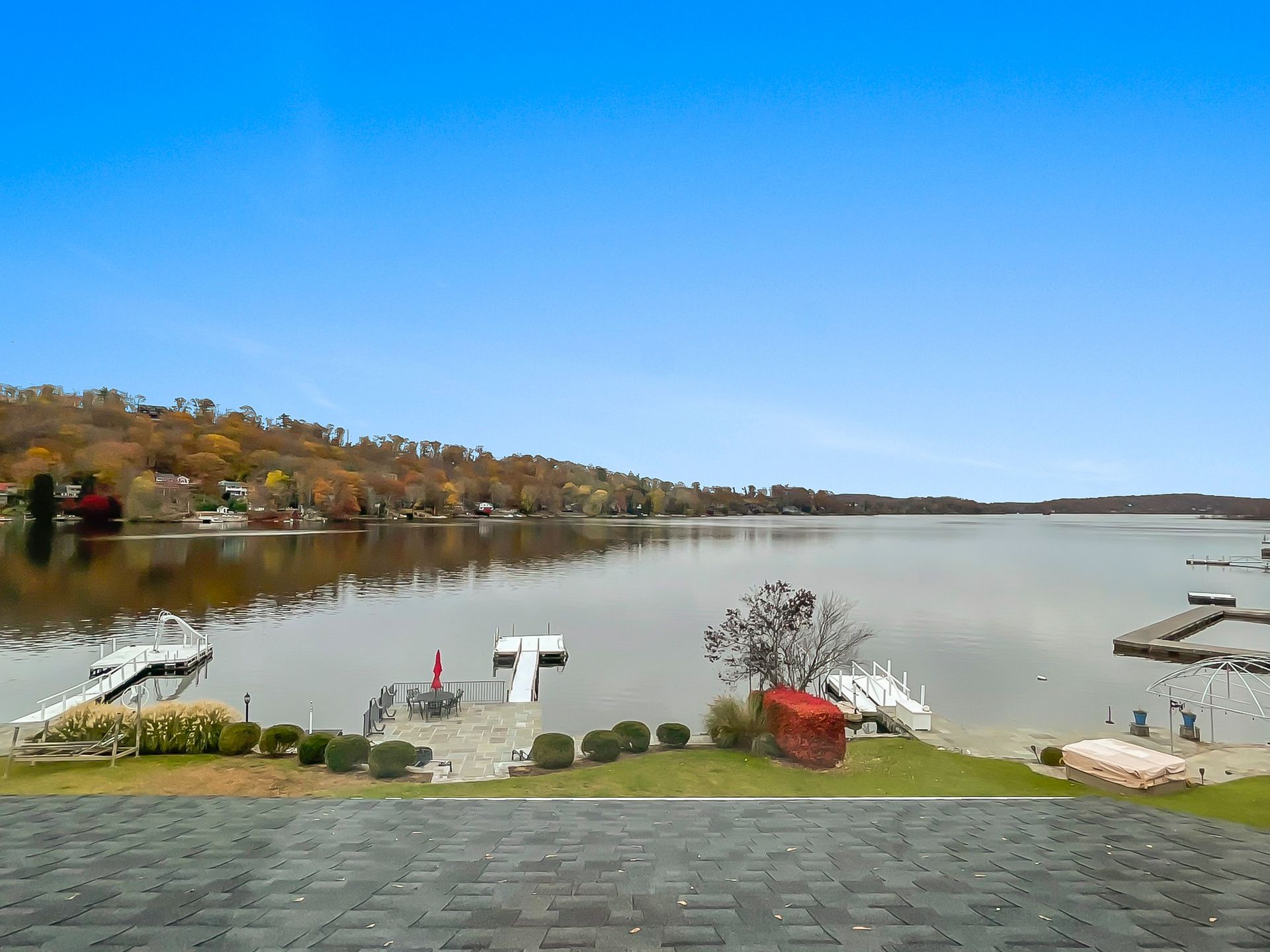 A scenic view of a calm lake from above, featuring wooden docks, green lawn, autumn trees, and a clear blue sky.