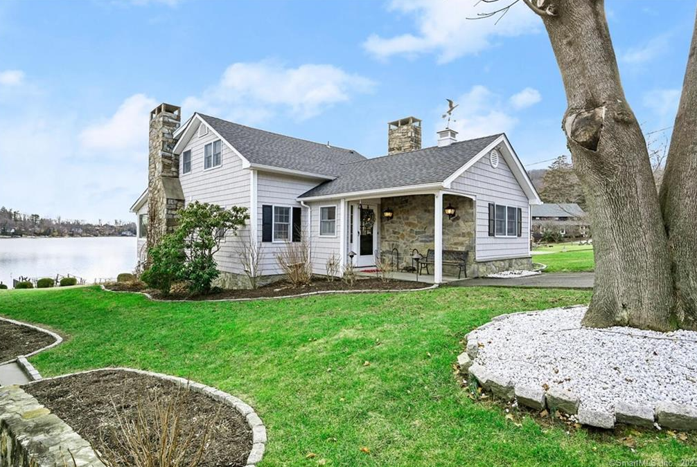 A light-colored house with stone chimneys and a covered porch, set on a green lawn near a calm lake.