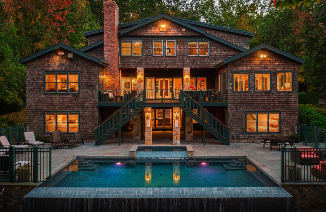A two-story brown brick house with an outdoor pool, stone patio, and wooden stairs at dusk.