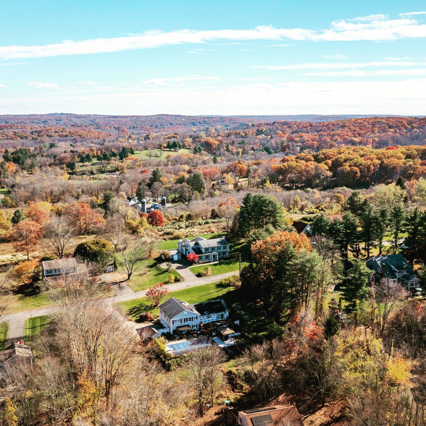 Aerial view of suburban houses nestled among autumn trees in a hilly, colorful landscape under a bright blue sky.