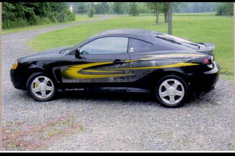 A black car with yellow stripes on the side is parked on gravel.