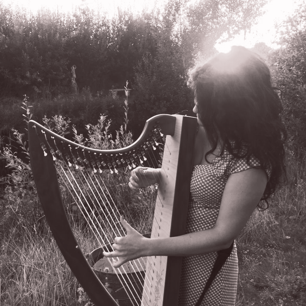 Jennette Roozeboom plays a harp in an outdoor, sunlit field. The image is in black and white.