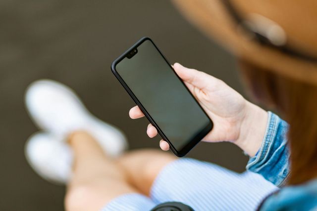 Pessoa segurando um smartphone preto com tela em branco, sentada do lado de fora.