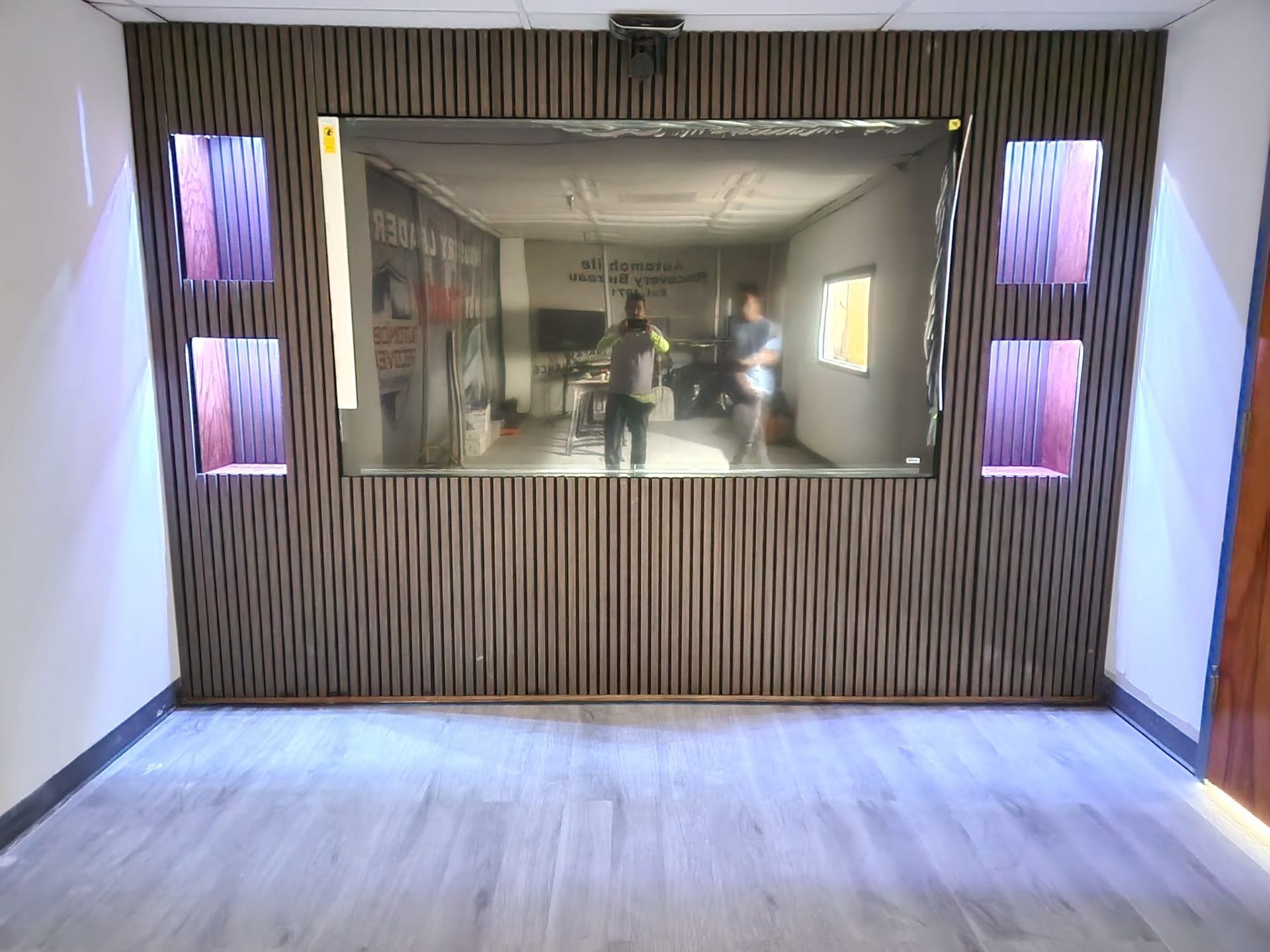 Room with a large mirror reflecting two people, wooden paneling, and square illuminated alcoves.