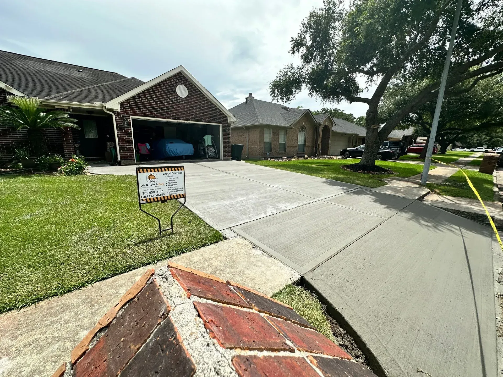 Residential street scene; house with garage, sign on lawn, concrete driveway and sidewalk, brick mailbox.