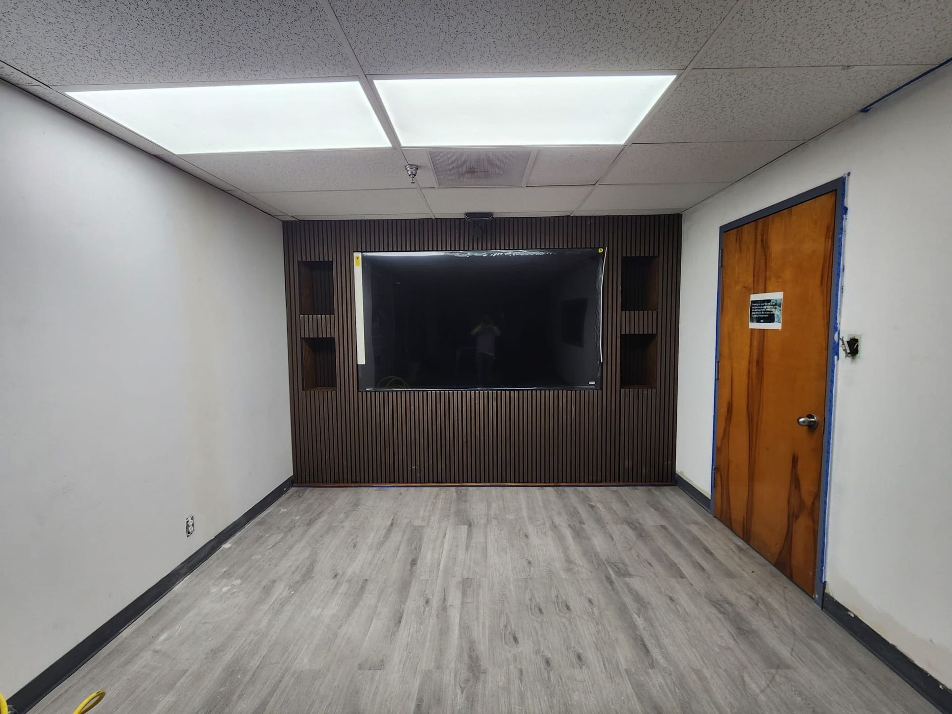 Empty room with large TV on brown draped wall, two shelves, wood door, and grey floor.