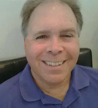 Man smiling, wearing a blue collared shirt. Indoor setting with a dark background.