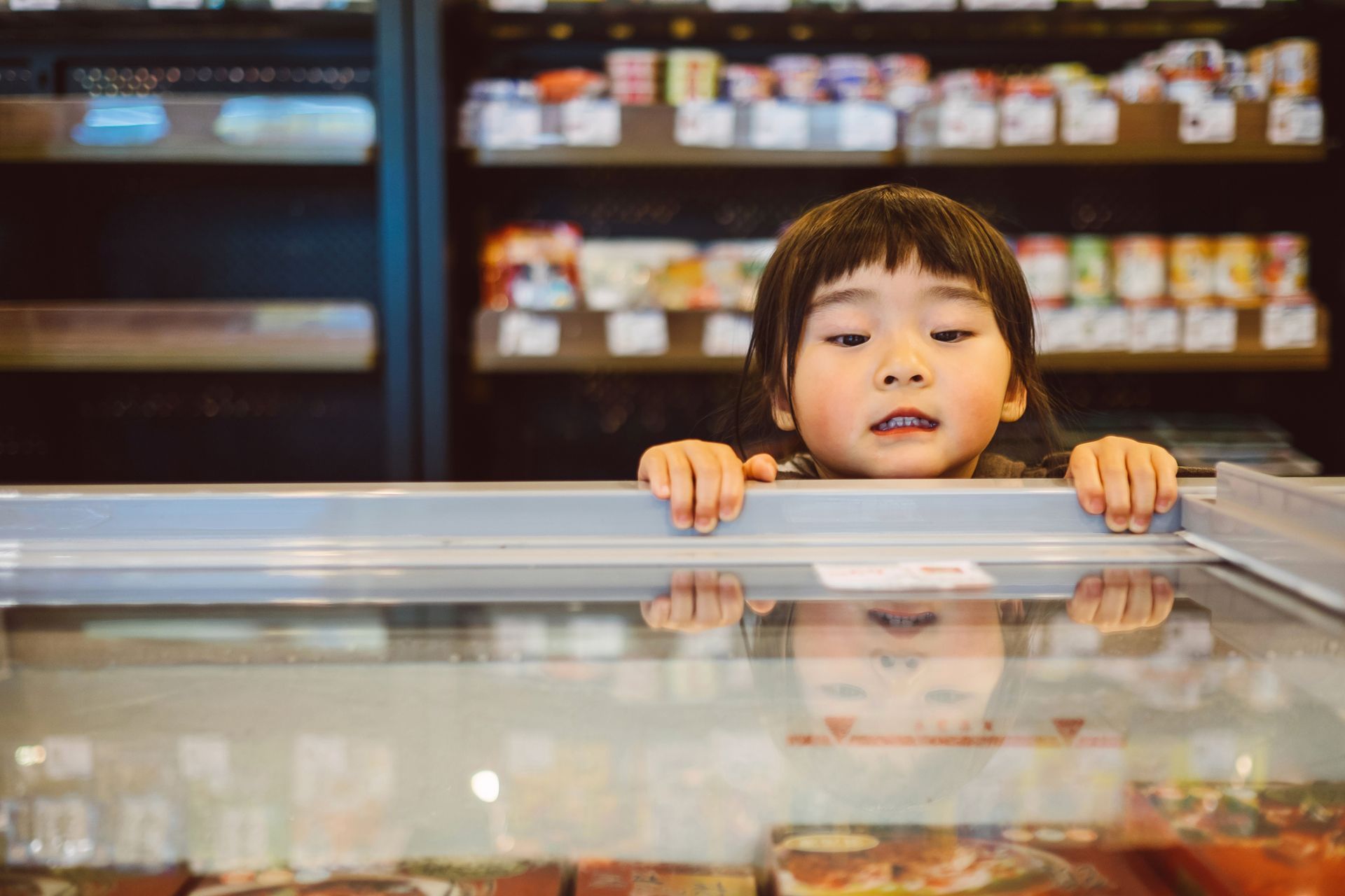 A young boy is peeking over a glass display case in a grocery store.