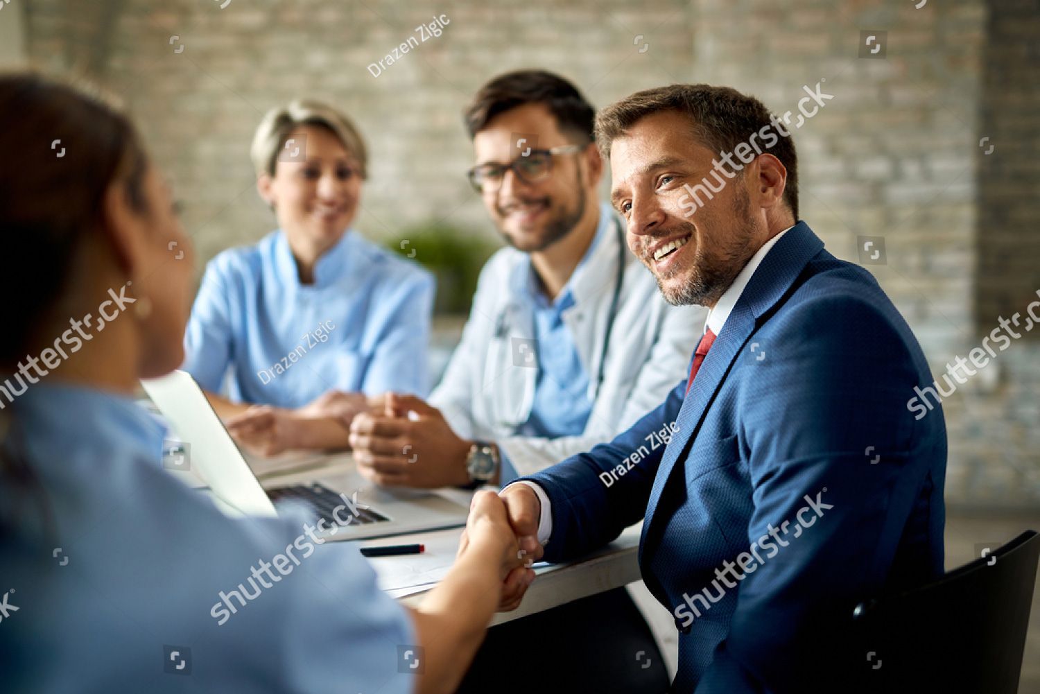 Man in suit shaking hands with a healthcare worker in a meeting with two other healthcare professionals, all smiling.