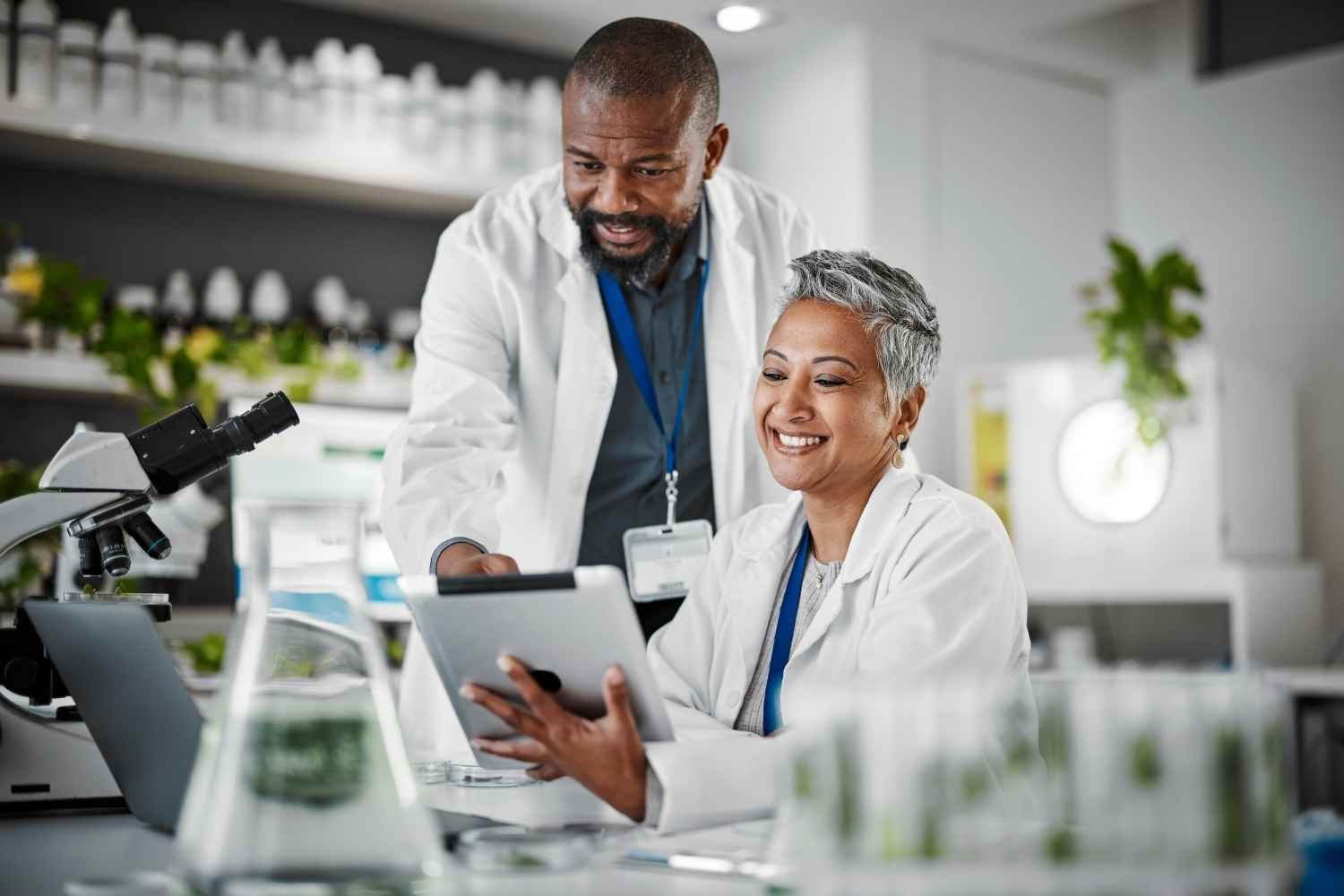 Two scientists in lab coats looking at a tablet, discussing results, with a lab setting background.