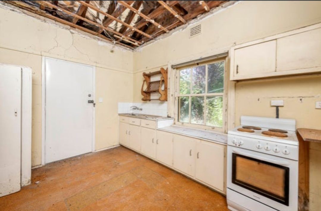 Old, Worn Kitchen With Exposed Ceiling. White Cabinets, Stove, and Door. Window and Sink Along One Wall  — Elimar Construction in Goulburn, NSW