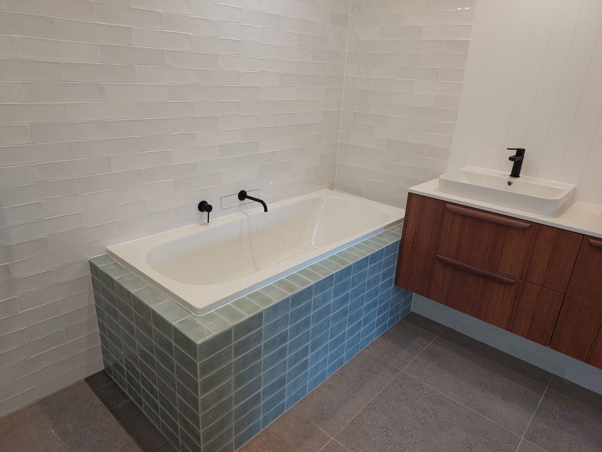 Bathroom With White Rectangular Tub, Surrounded by Blue-green Brick. White Walls, Wood Vanity With Sink, Black Fixtures — Elimar Construction in Goulburn, NSW