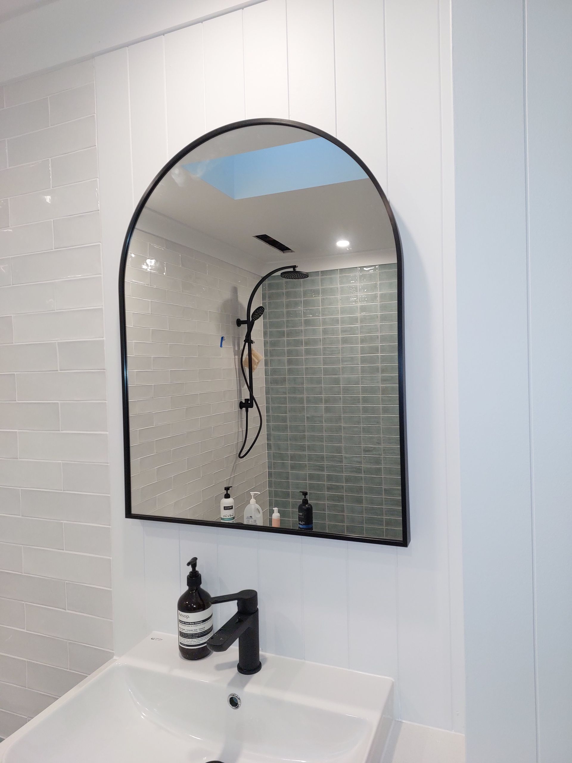 Bathroom With Arched Black-framed Mirror, White Sink, and Showerhead Visible in Reflection; White and Blue-tiled Walls — Elimar Construction in Goulburn, NSW
