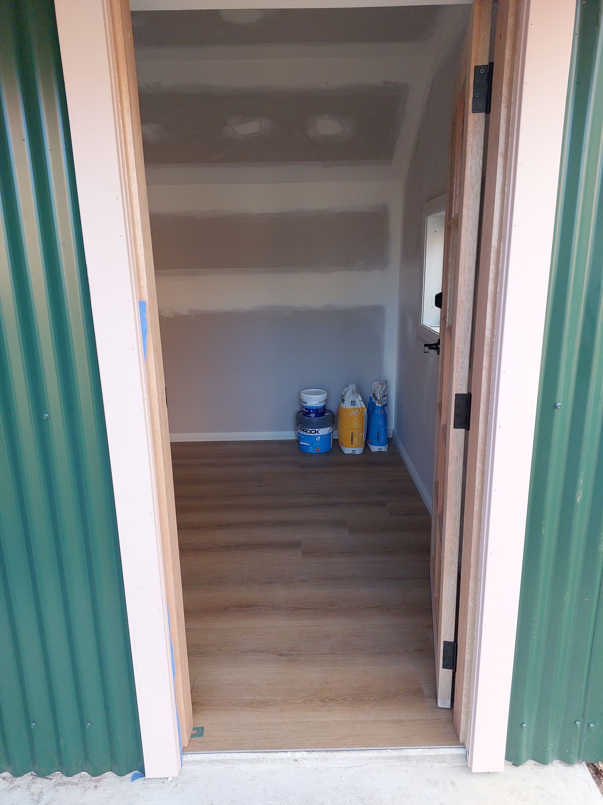 Open Doorway Into a Small Storage Room With Paint Cans and Wood-look Flooring. Green Corrugated Exterior — Elimar Construction in Goulburn, NSW