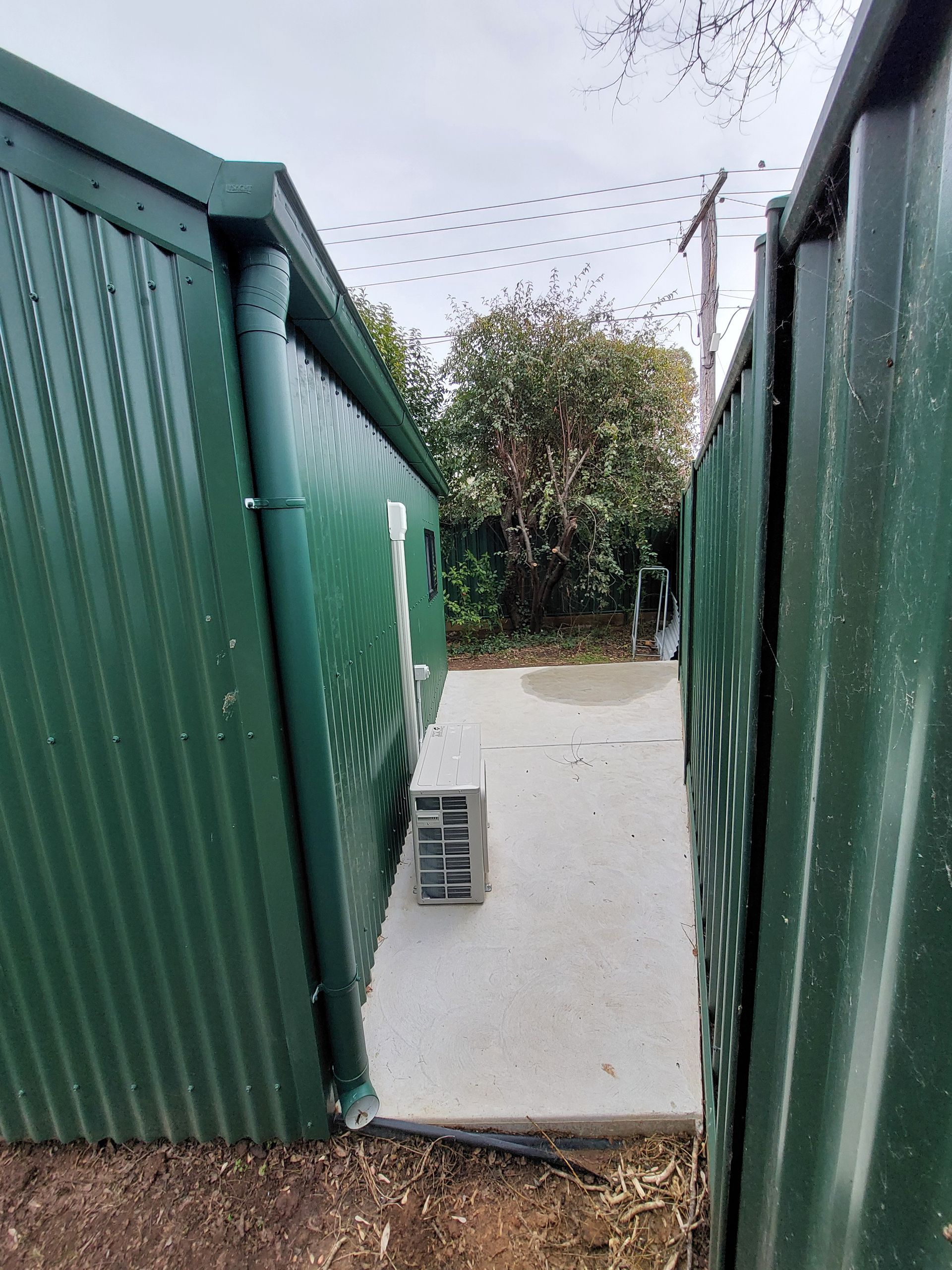 Green Corrugated Metal Sheds With Concrete Walkway, Small Air Conditioning Unit, and Foliage in Background — Elimar Construction in Goulburn, NSW
