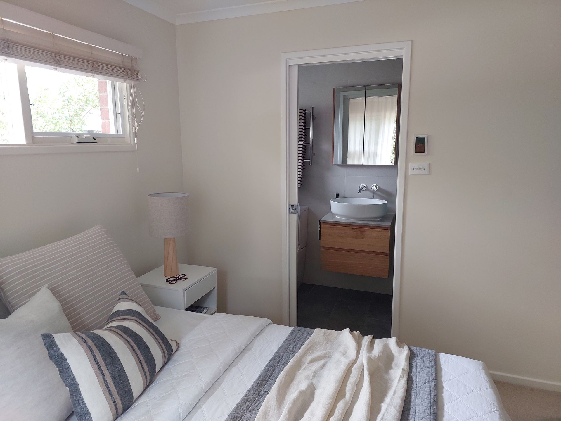 Bedroom With Bed, Nightstand, Window, and Doorway Leading to a Bathroom With Vanity and Mirror — Elimar Construction in Goulburn, NSW