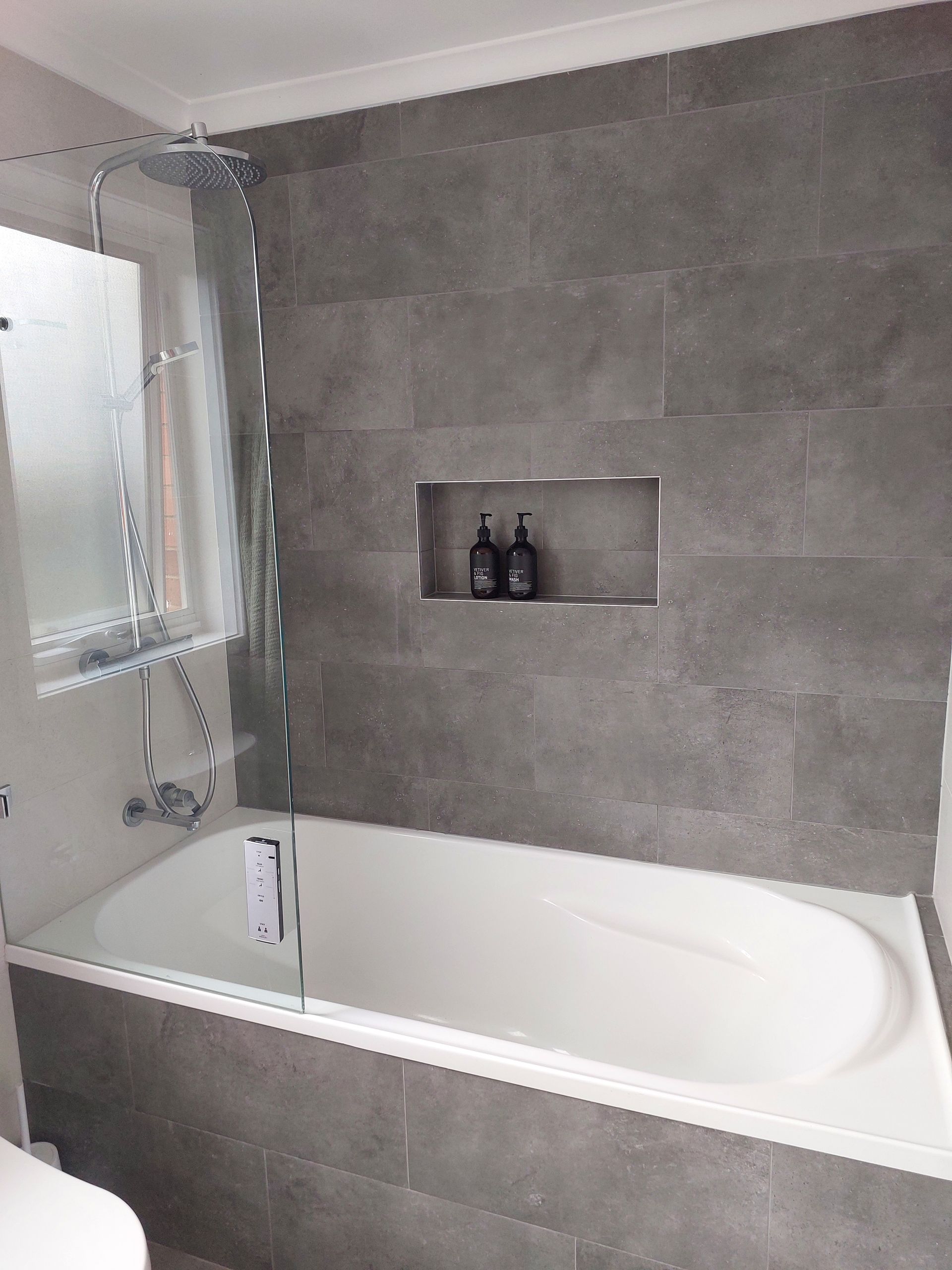 Bathroom With Gray Tiled Walls, Bathtub, Glass Shower Screen, and Recessed Shelf With Toiletries — Elimar Construction in Goulburn, NSW