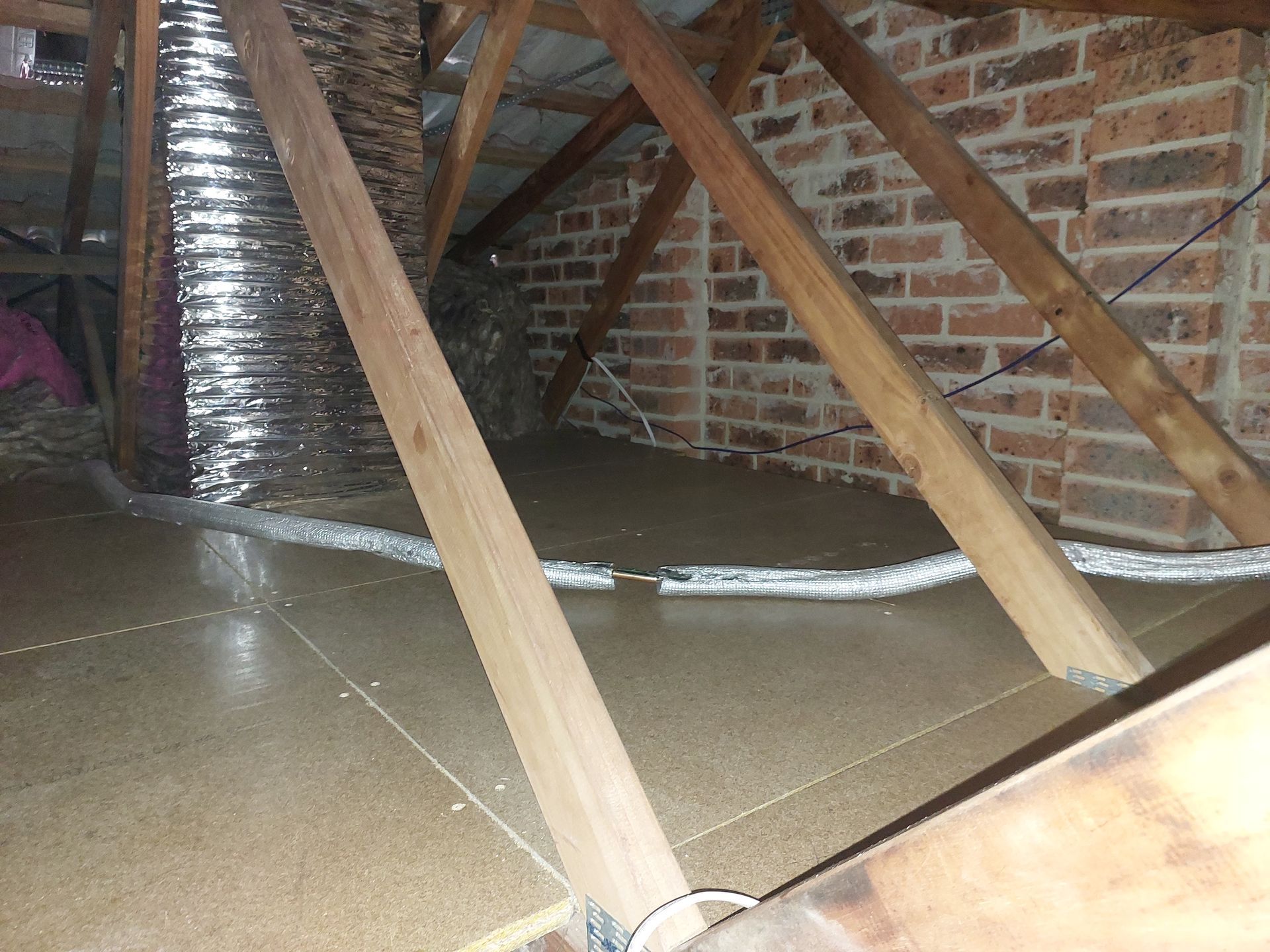 Attic Interior With Wooden Rafters, Brick Wall, and Flexible Ductwork. Brown Insulation Covers the Floor — Elimar Construction in Goulburn, NSW