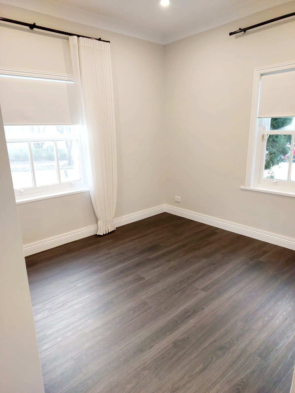 Empty Room With Dark Wood-look Floor, White Walls, Two Windows With Roller Blinds, and White Curtains  — Elimar Construction in Goulburn, NSW