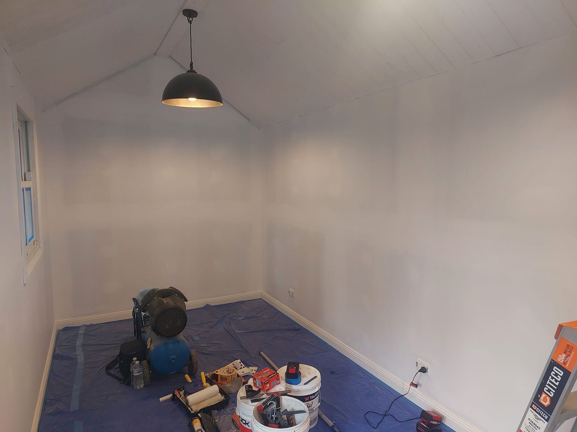 Interior of a Room Painted White, With Blue Tarp on the Floor, and Painting Supplies. a Light Fixture Hangs From the Ceiling — Elimar Construction in Goulburn, NSW