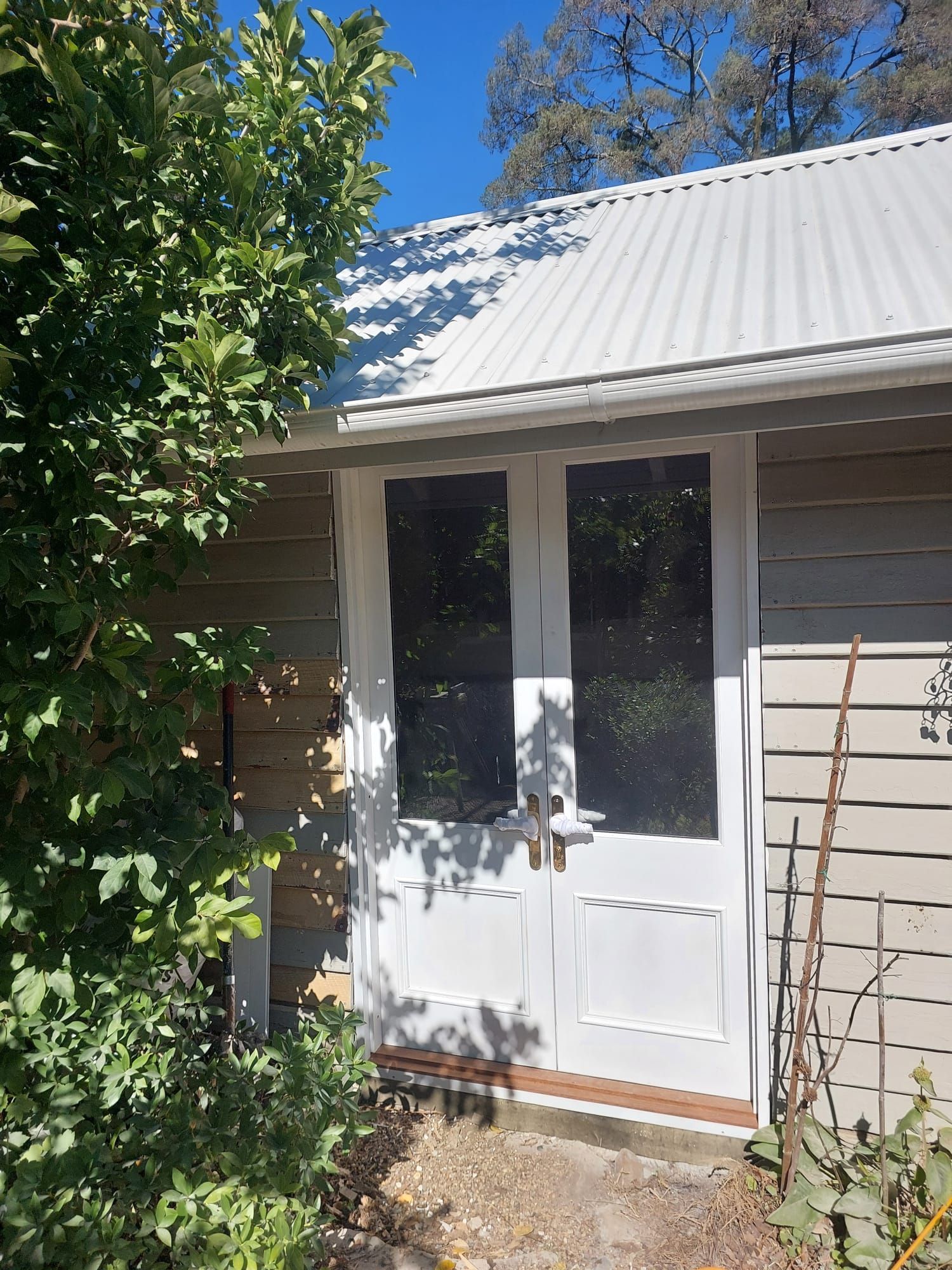 White Double Doors on a Weathered Wooden Building With Corrugated Metal Roof, Partly Obscured by Foliage — Elimar Construction in Goulburn, NSW
