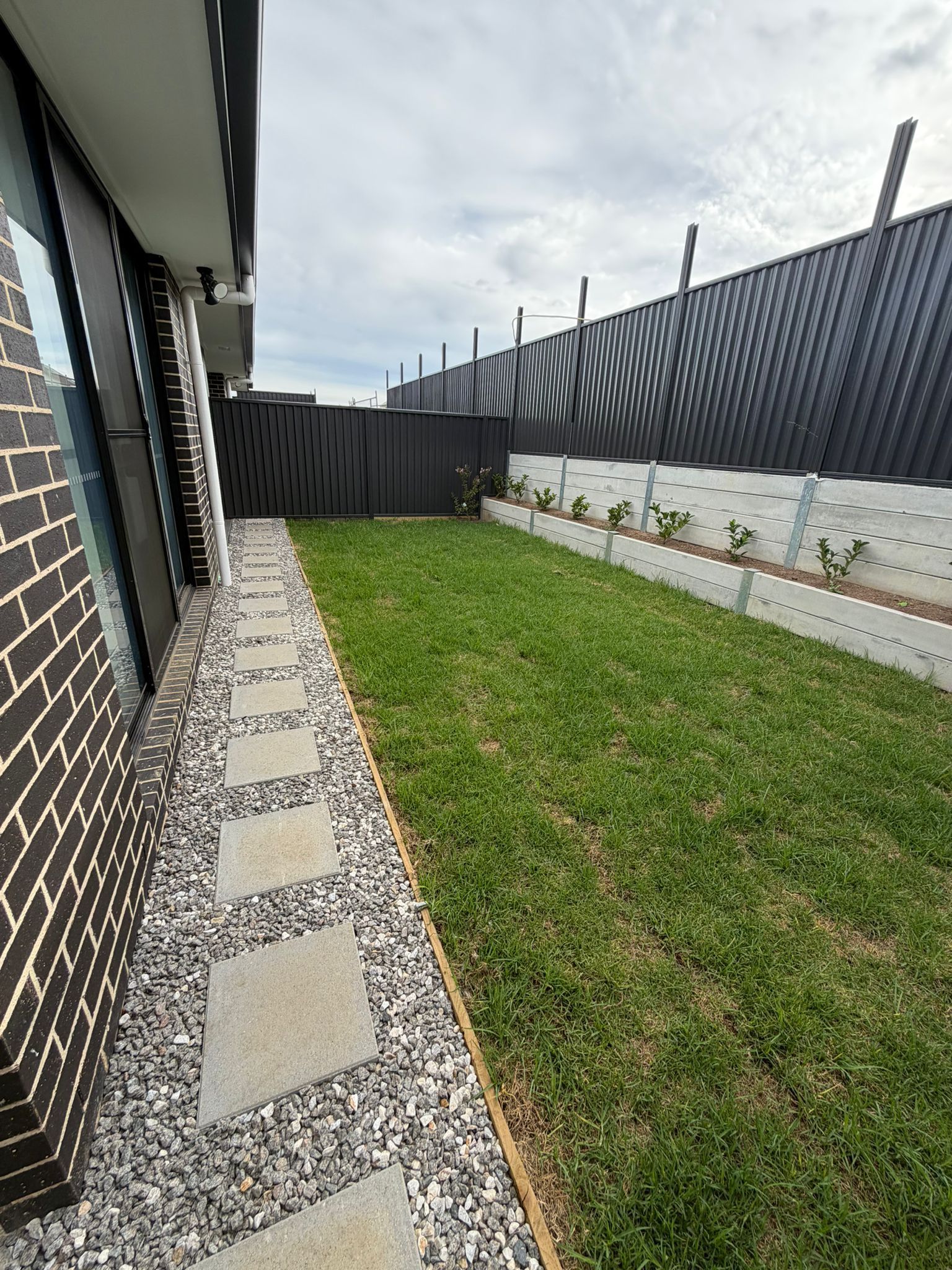 Narrow side yard with grass, stepping stones, and a house wall beside a tall fenced perimeter — Elimar Construction in Goulburn, NSW