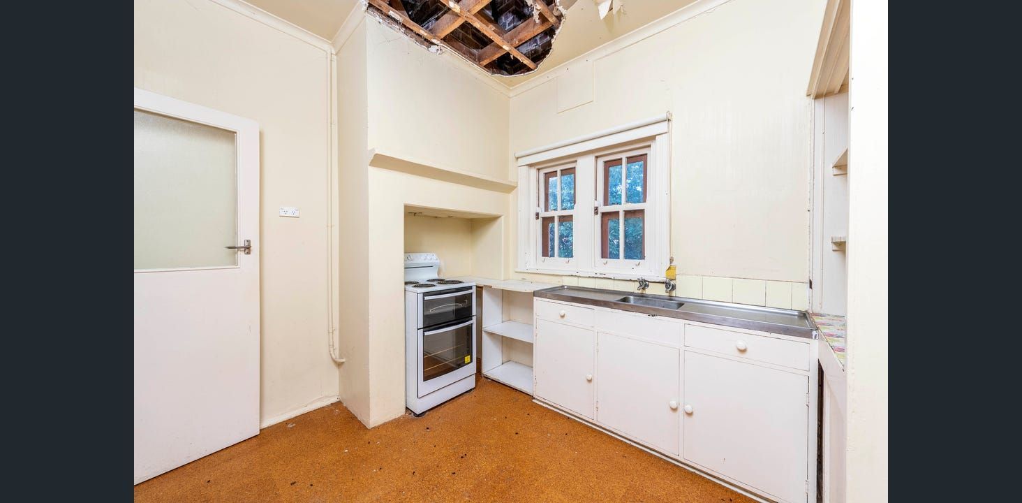 Small, Dated Kitchen With White Cabinets, a Stove, and a Window. the Ceiling is Damaged  — Elimar Construction in Goulburn, NSW