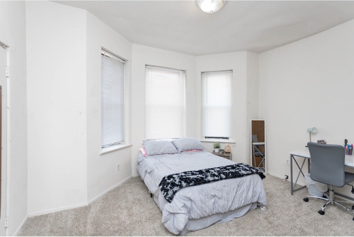 Bedroom with bed, desk, and three windows with blinds. White walls and gray carpet.
