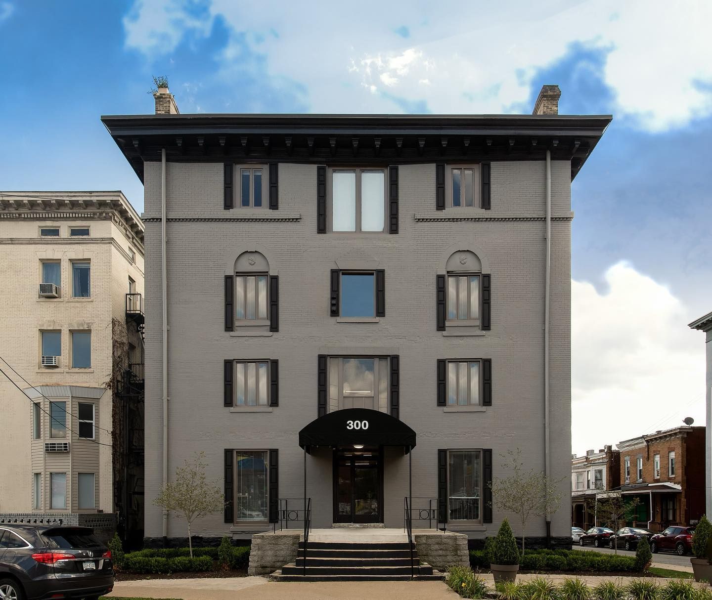 Gray brick building with black trim and awning; trees and parked cars in front.