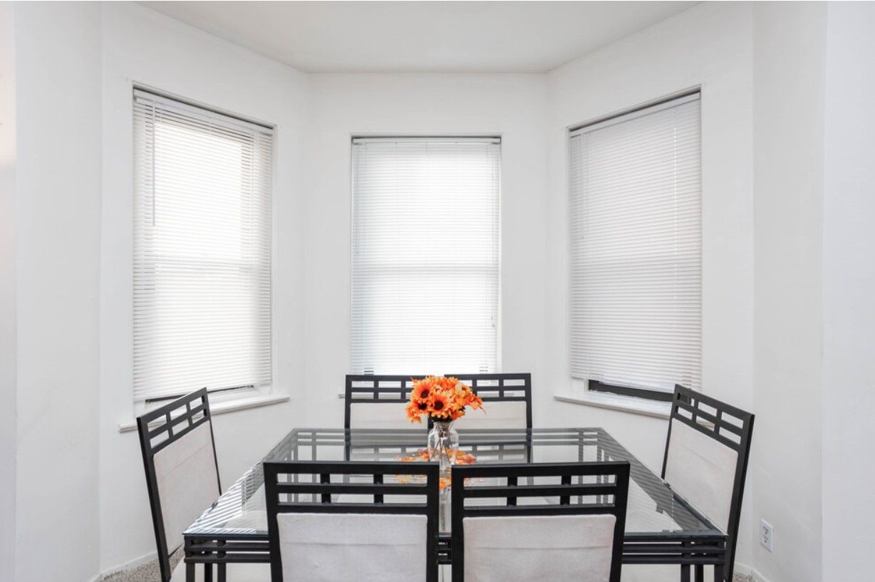 Dining room with square glass table, black chairs, and bay windows with blinds. Orange flowers in center.
