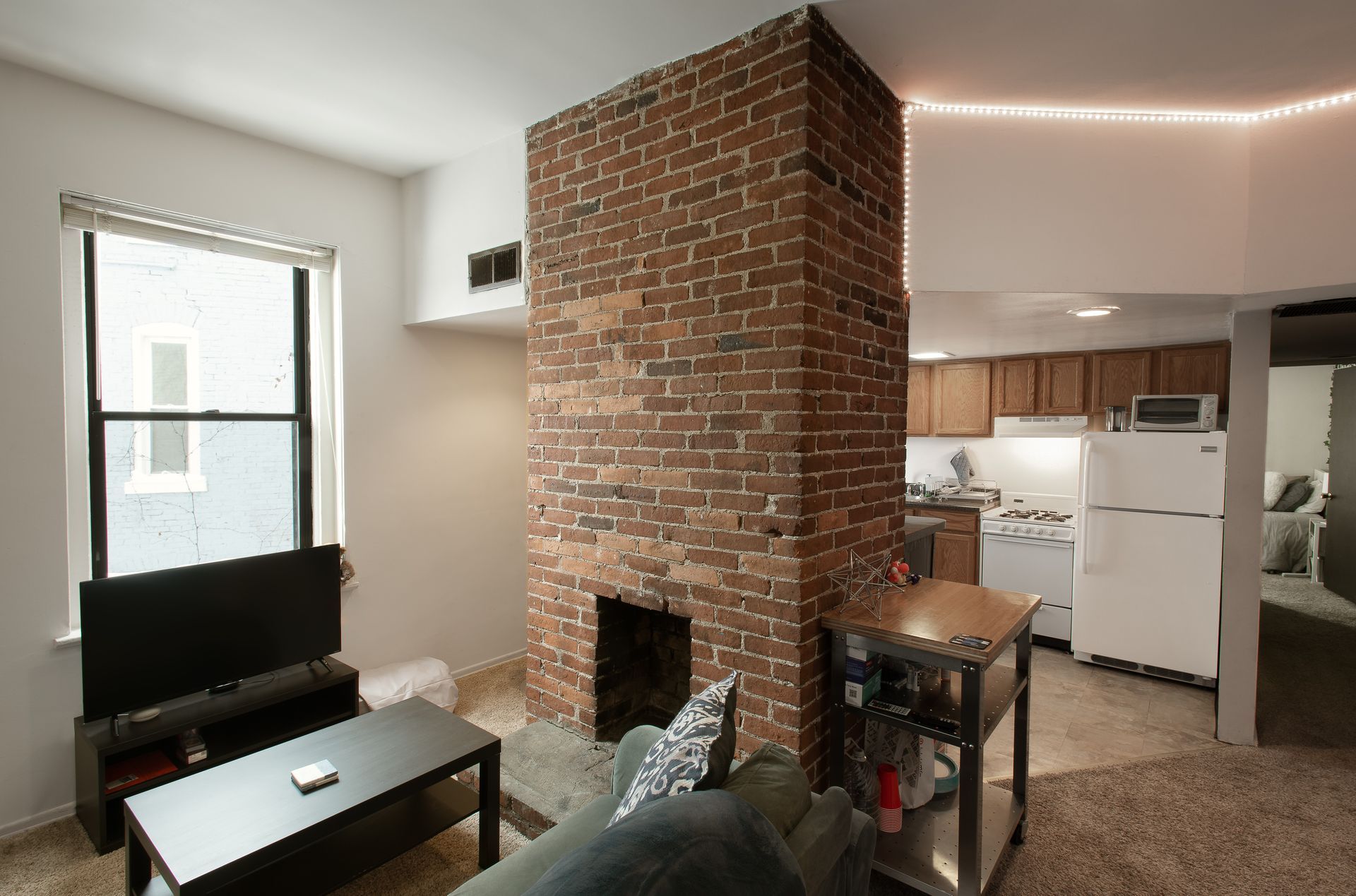 Living room with exposed brick chimney, window, TV, sofa, and open kitchen.