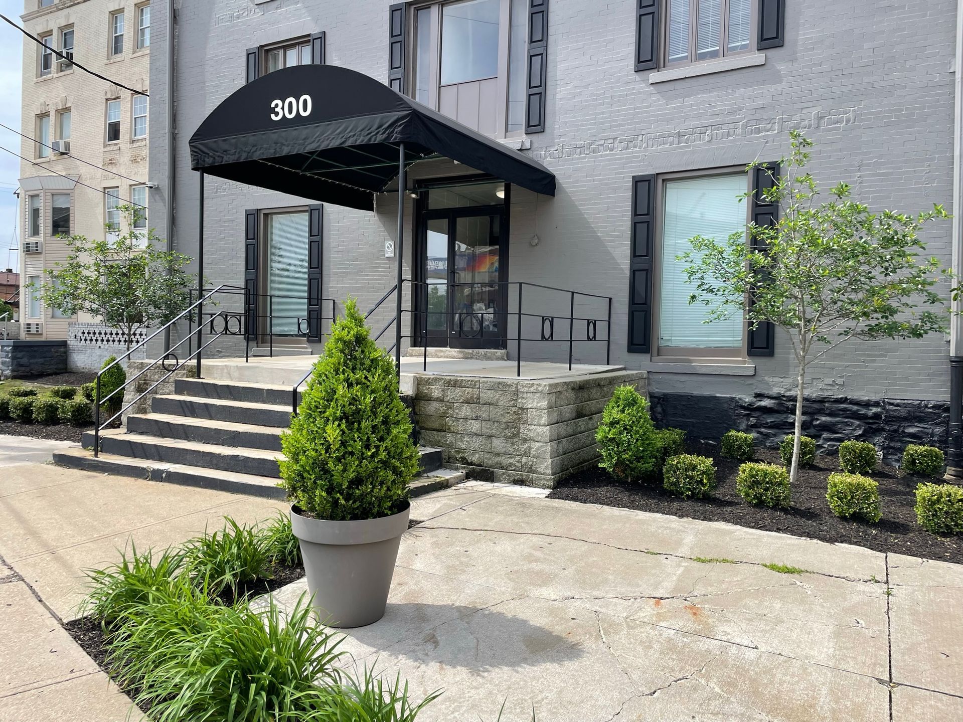 Gray building entrance with black awning, steps, and a large potted evergreen in front.