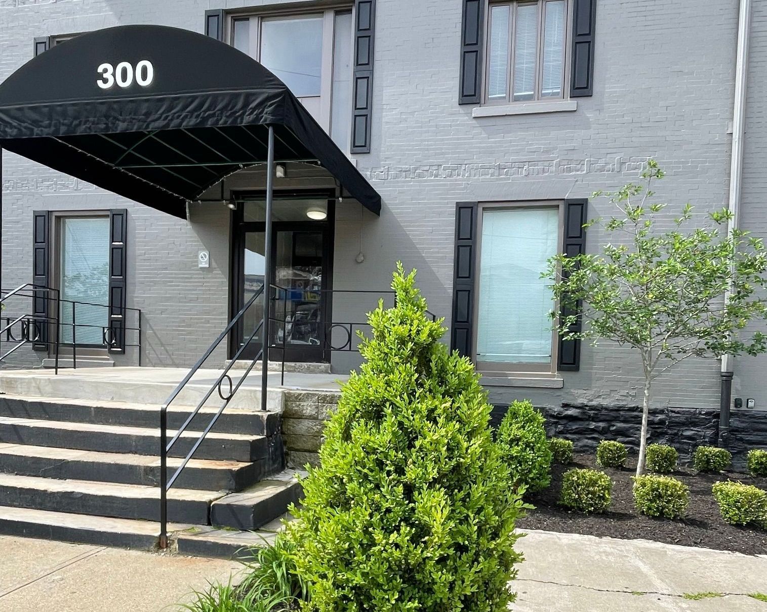 Gray building with black awning and shutters. Steps lead to the entrance.