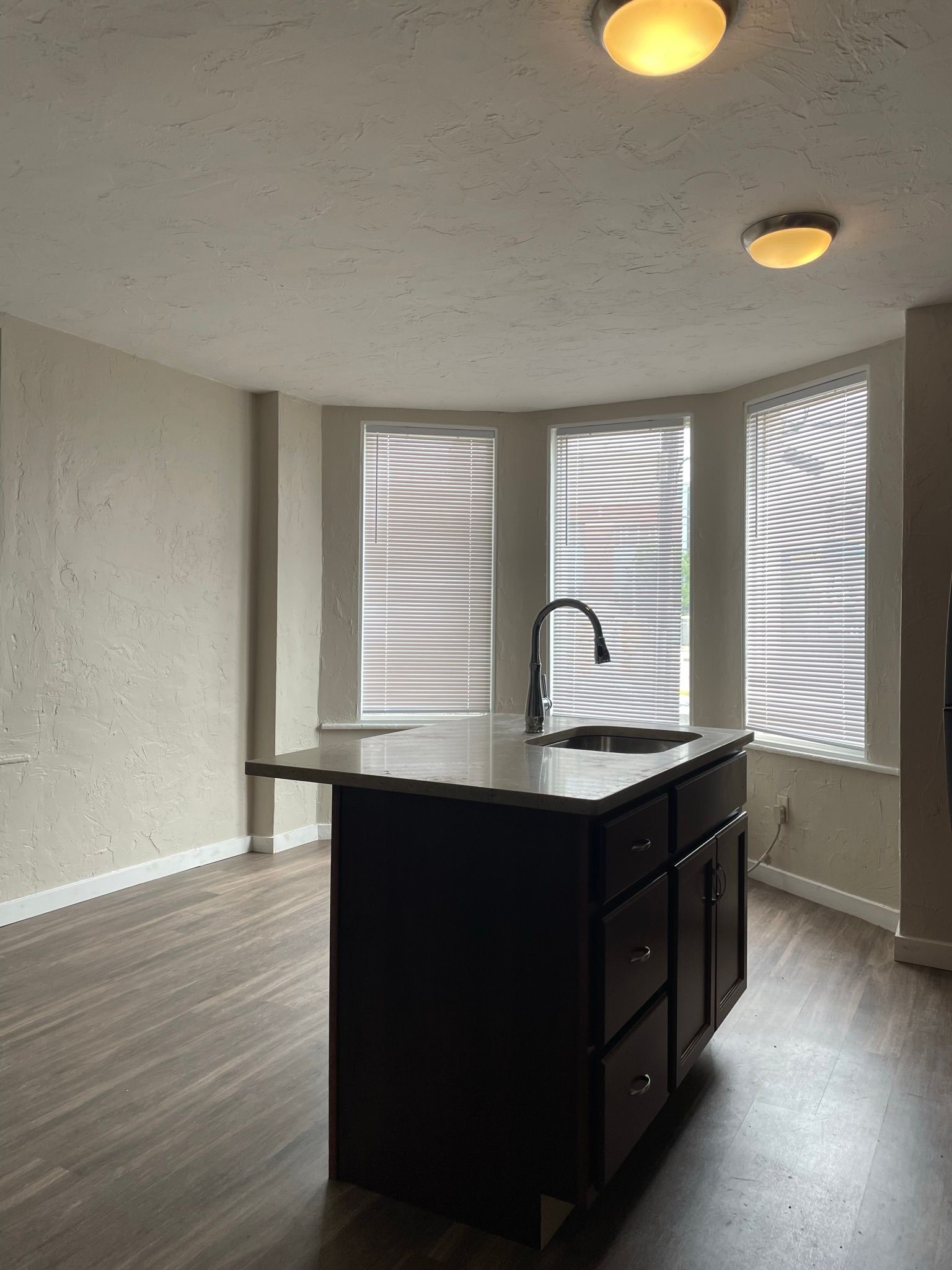 Kitchen with dark island, sink, and bay window with blinds; wood floors.