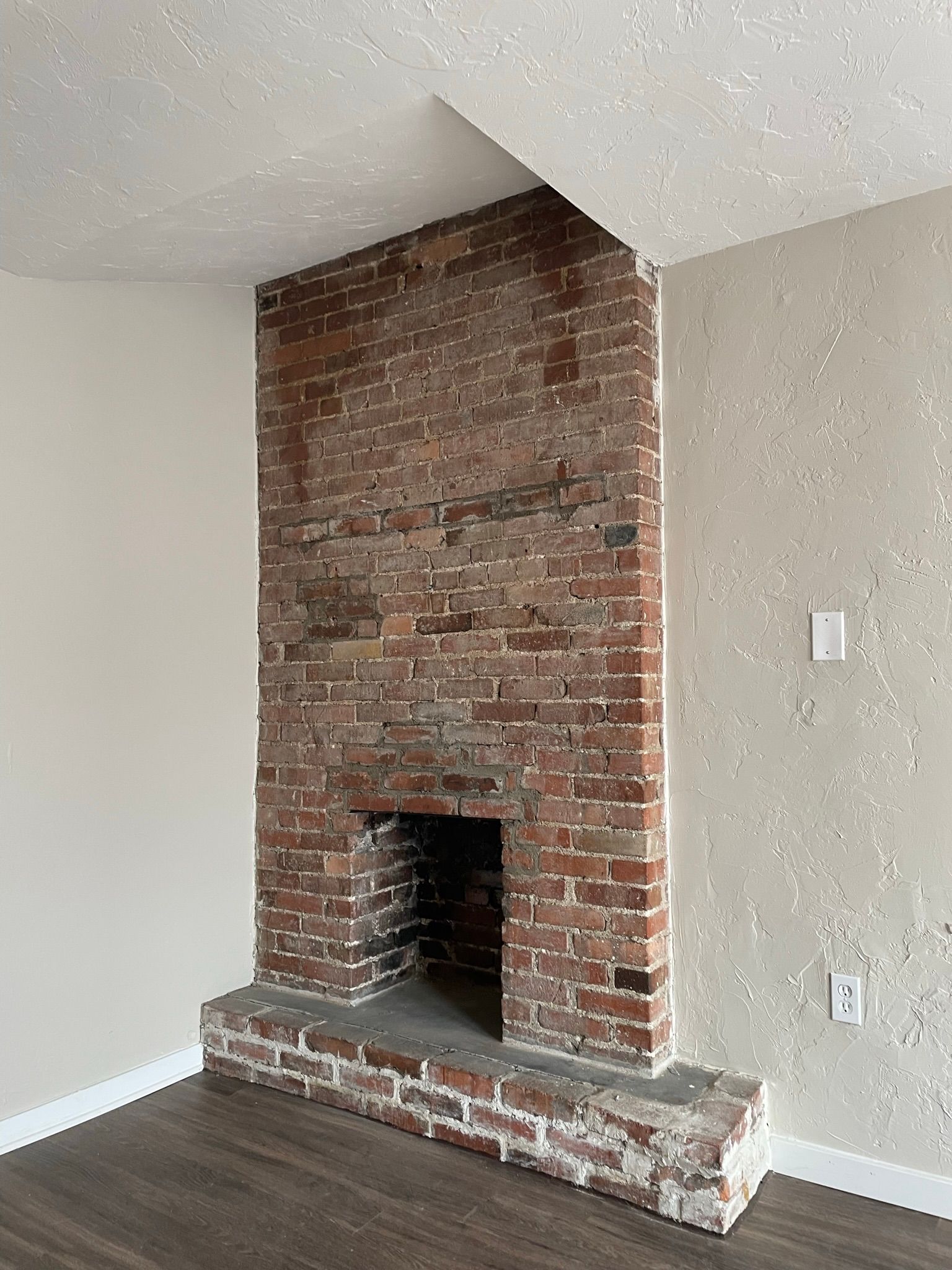 Brick fireplace in corner of room with wood floors and off-white walls.