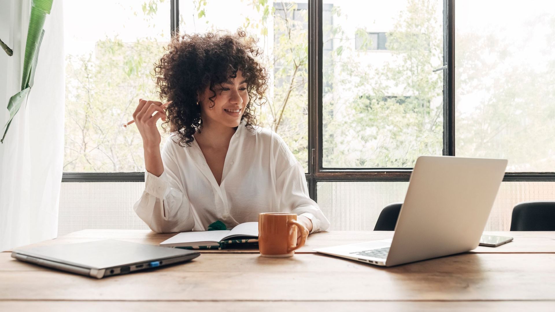 Woman with curly hair smiles, working on laptop at a wooden desk near a window, with a mug and notepad.
