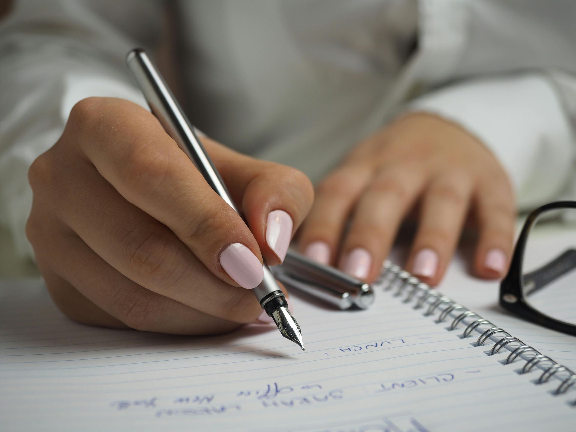 Woman's hands writing with a silver pen in a notebook, light pink nail polish, glasses in the background.