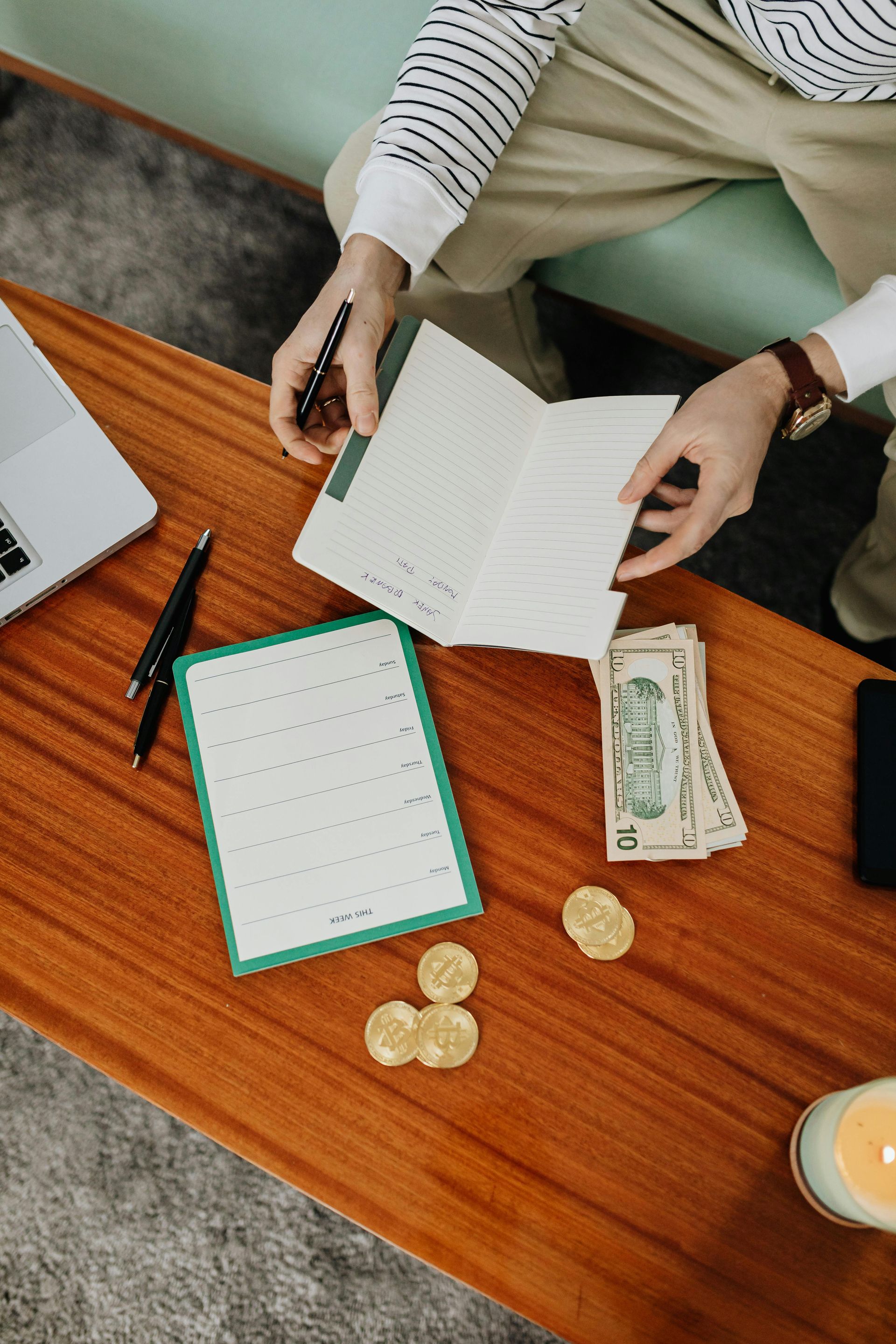 Person reviewing finances at a table with notebook, cash, coins, laptop, and candle.
