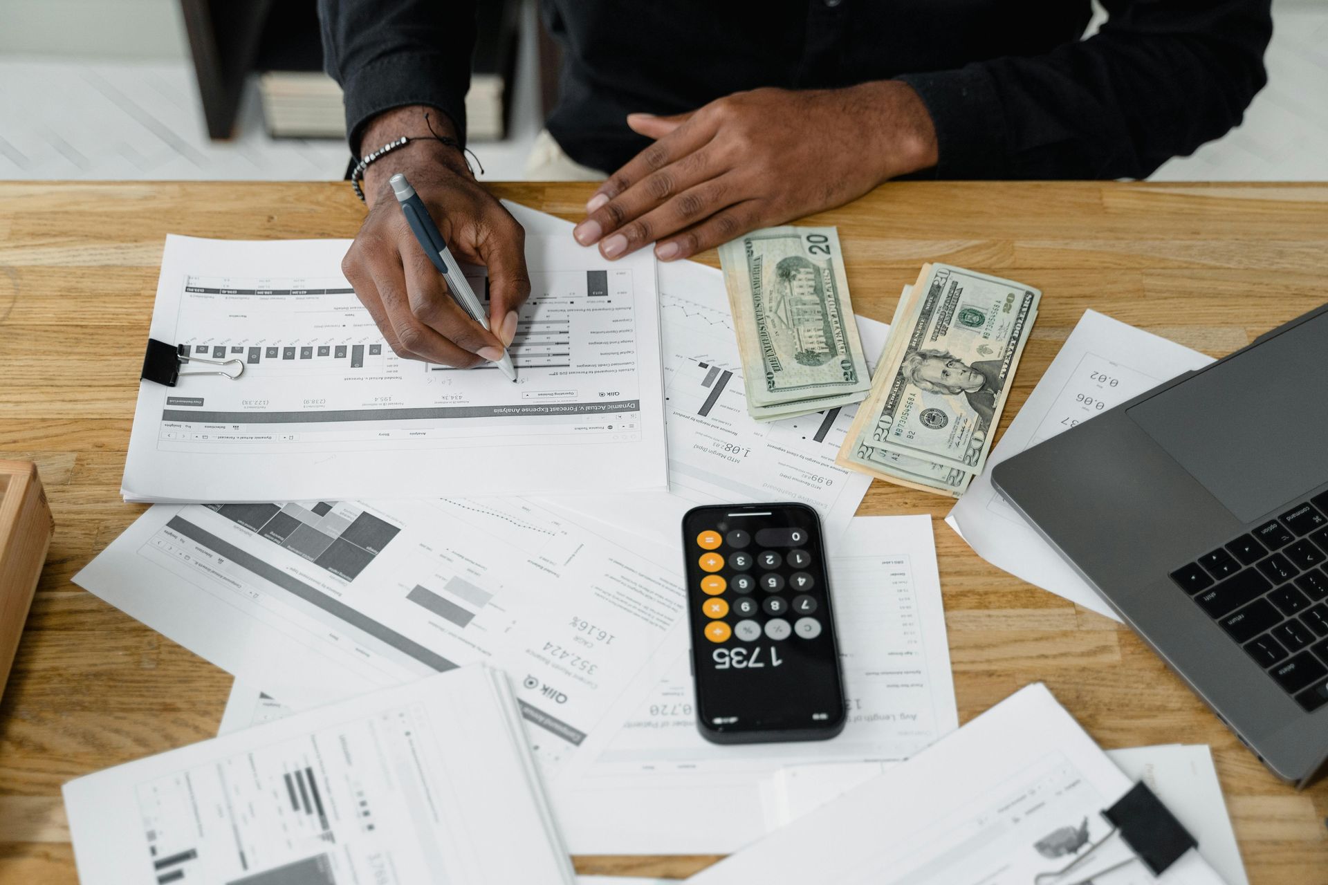Person with pen calculating finances on desk with cash, calculator, and laptop.