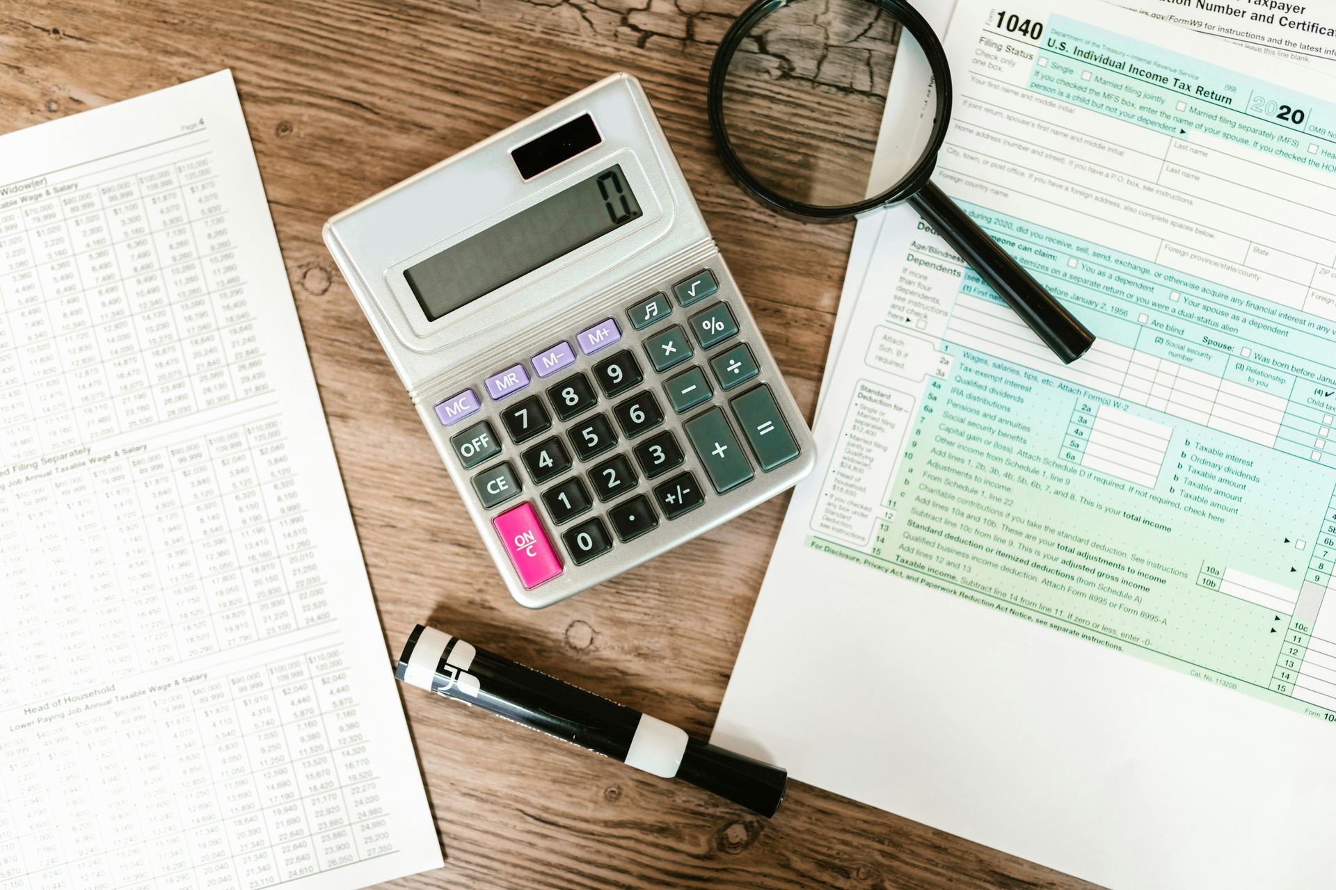 Calculator, tax forms, magnifying glass, and pen on a wooden desk, suggesting tax preparation.
