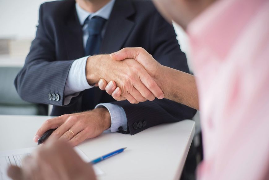 Two men in a business setting shake hands over a desk; one in a suit, the other in a pink shirt.
